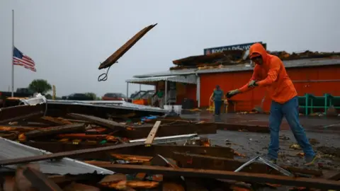 reuters a man in a bright orange jacket tosses a plank of wood onto a pile during a hurricane clean up. an american flag at half mast is behind him, as is on orange building with a damaged roof and a sign on it. 